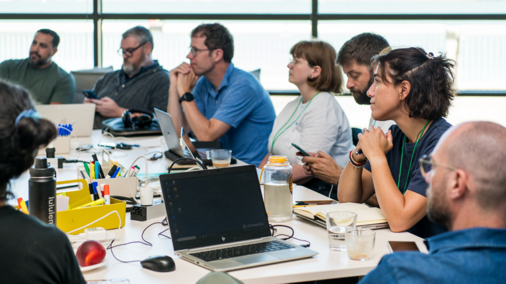 The image shows a group of individuals seated around a table during a workshop or meeting. They are focused on discussions or presentations, with some taking notes, using laptops, or checking their phones. The table is cluttered with laptops, notebooks, markers, water bottles, and other office supplies, indicating an active and collaborative environment. The background reveals a bright room with large windows, suggesting a modern, open setting.