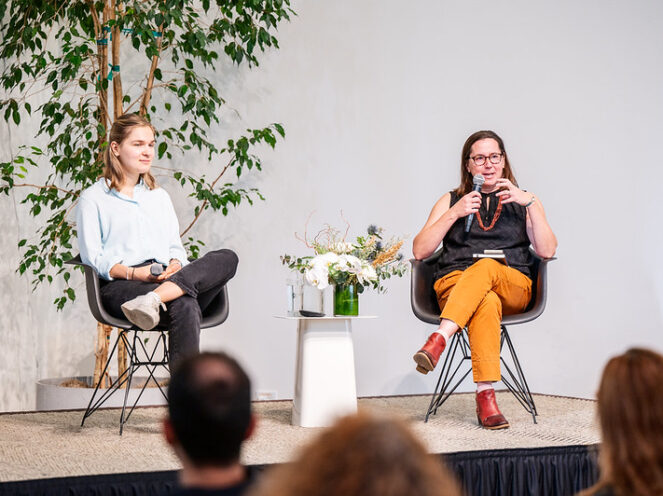 Two women seated on a stage in modern black chairs, engaging in a panel discussion or dialogue. One woman, dressed in a blue blouse and black jeans, listens attentively with a relaxed posture, while the other, wearing a black top, orange pants, and red shoes, speaks into a microphone. Between them is a small white table adorned with a floral arrangement and a glass of water. The background features a minimalist design with green foliage, creating a calm and inviting atmosphere. Audience members' heads are visible in the foreground, indicating an engaged audience.