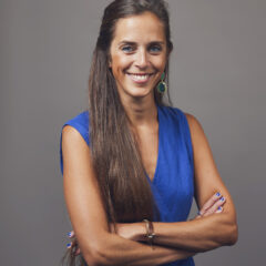 This is a professional portrait of a woman wearing a vibrant blue dress. She has long brown hair, styled neatly, and is accessorized with statement green earrings and bracelets. She is smiling warmly at the camera, standing confidently with her arms crossed. The neutral gray background draws attention to her engaging expression and attire, making the image suitable for professional or promotional contexts.