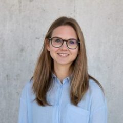 The image portrays a young woman with long, light brown hair, wearing glasses and a light blue shirt. She is smiling and standing in front of a plain, neutral background, giving off a professional yet approachable vibe.