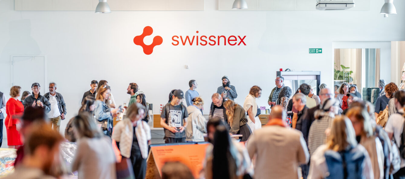 A vibrant gathering at Swissnex featuring a crowd of attendees exploring exhibits and networking in a bright, modern space. The Swissnex logo is prominently displayed on the wall, emphasizing the innovative and collaborative atmosphere. Attendees engage with displays, converse in small groups, and move throughout the open area, which is illuminated by natural light and modern overhead fixtures. The event radiates energy, creativity, and a sense of community, showcasing the spirit of innovation and exchange.
