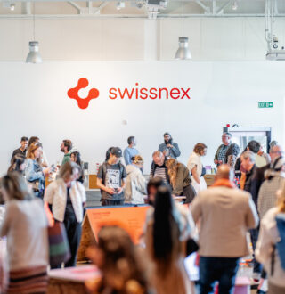A vibrant gathering at Swissnex featuring a crowd of attendees exploring exhibits and networking in a bright, modern space. The Swissnex logo is prominently displayed on the wall, emphasizing the innovative and collaborative atmosphere. Attendees engage with displays, converse in small groups, and move throughout the open area, which is illuminated by natural light and modern overhead fixtures. The event radiates energy, creativity, and a sense of community, showcasing the spirit of innovation and exchange.