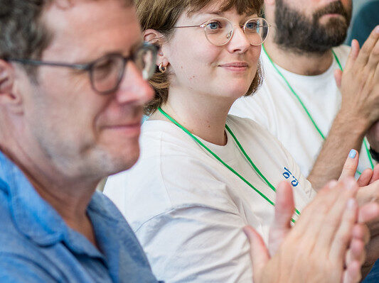 A candid moment during a workshop featuring three participants clapping, indicating appreciation or acknowledgment. The man in the foreground wears a blue shirt and glasses, while the woman in the middle, wearing a white shirt, glasses, and a green lanyard, looks directly at the camera with a slight smile. Another man with a beard, seated in the background, also joins in the applause. The setting is bright and professional, with a sense of engagement and focus on the event's activities.