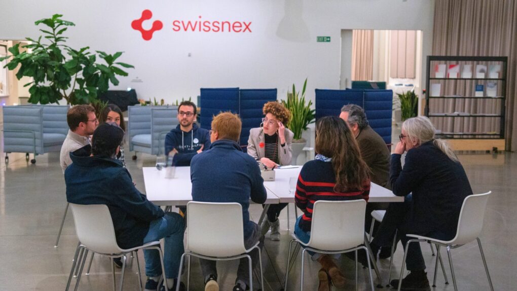 A group of people engaged in a focused discussion around a rectangular table in a modern, well-lit indoor space. The backdrop features the