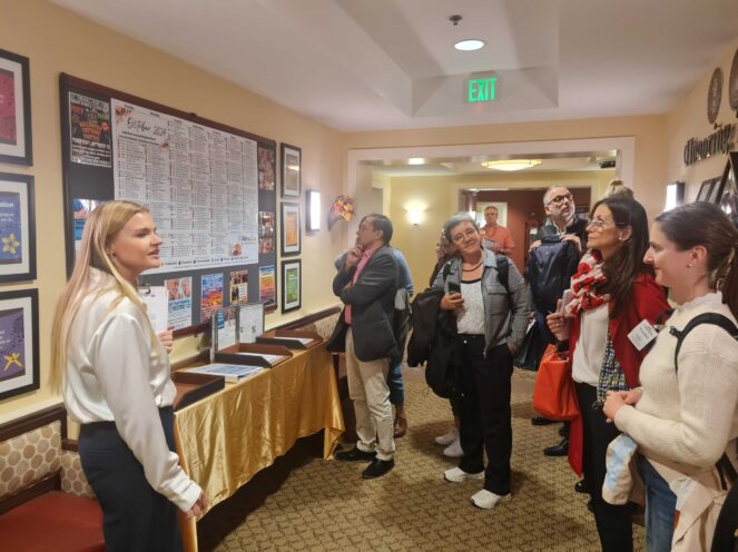 A guide speaking to a group of people in a hallway decorated with a bulletin board and framed posters. The attendees are engaged, standing in a small group, and listening attentively.