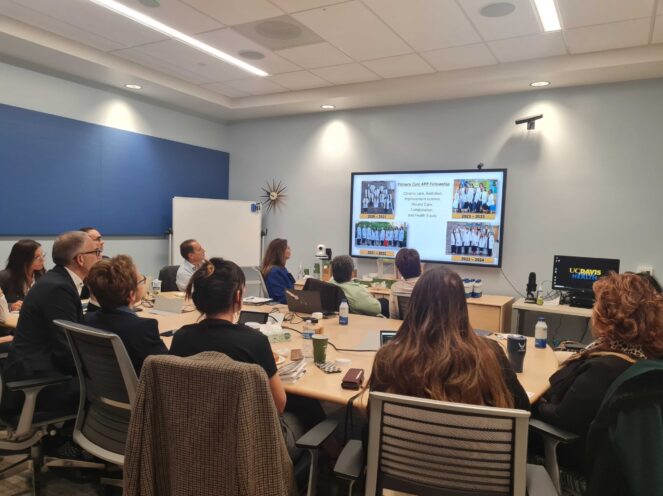 A group of individuals seated around a large table in a conference room, viewing a presentation displayed on a screen.