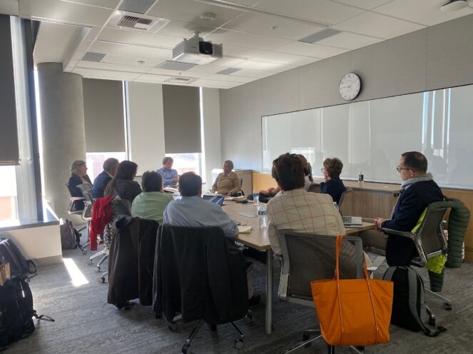 A team of people seated around a conference table in a well-lit room. A clock is visible on the wall along with large windows. Bags and coats are placed on the back of chairs.