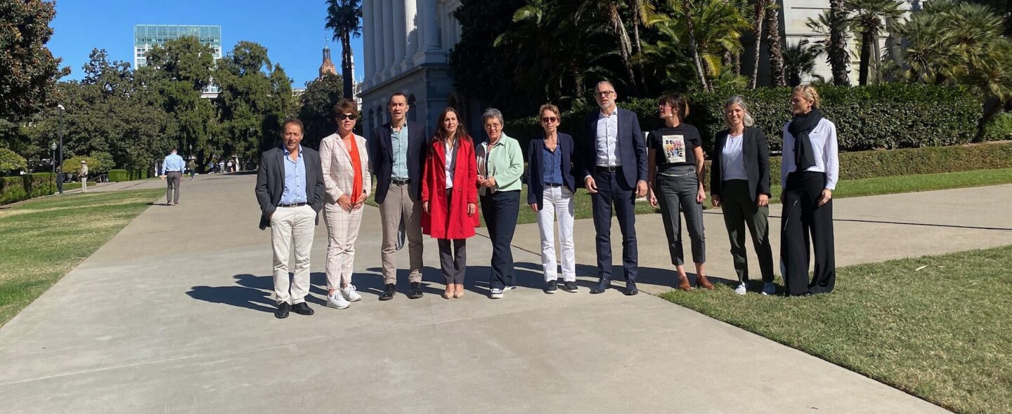 A group of ten people posing for a photo outdoors on a sunny day in front of a white neoclassical building surrounded by palm trees and lush greenery. The individuals are standing on a paved walkway, dressed in a mix of formal and casual attire, and the background shows a clear blue sky and distant buildings.
