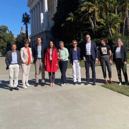 A group of ten people posing for a photo outdoors on a sunny day in front of a white neoclassical building surrounded by palm trees and lush greenery. The individuals are standing on a paved walkway, dressed in a mix of formal and casual attire, and the background shows a clear blue sky and distant buildings.