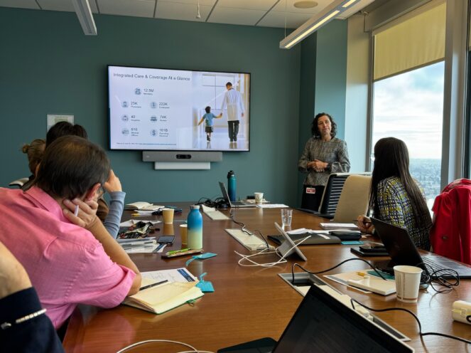 A group of attendees seated at a conference table in a modern meeting space with large windows. A presenter is speaking in front of a large screen displaying a presentation slide. Laptops, notebooks, and other materials are spread across the table.